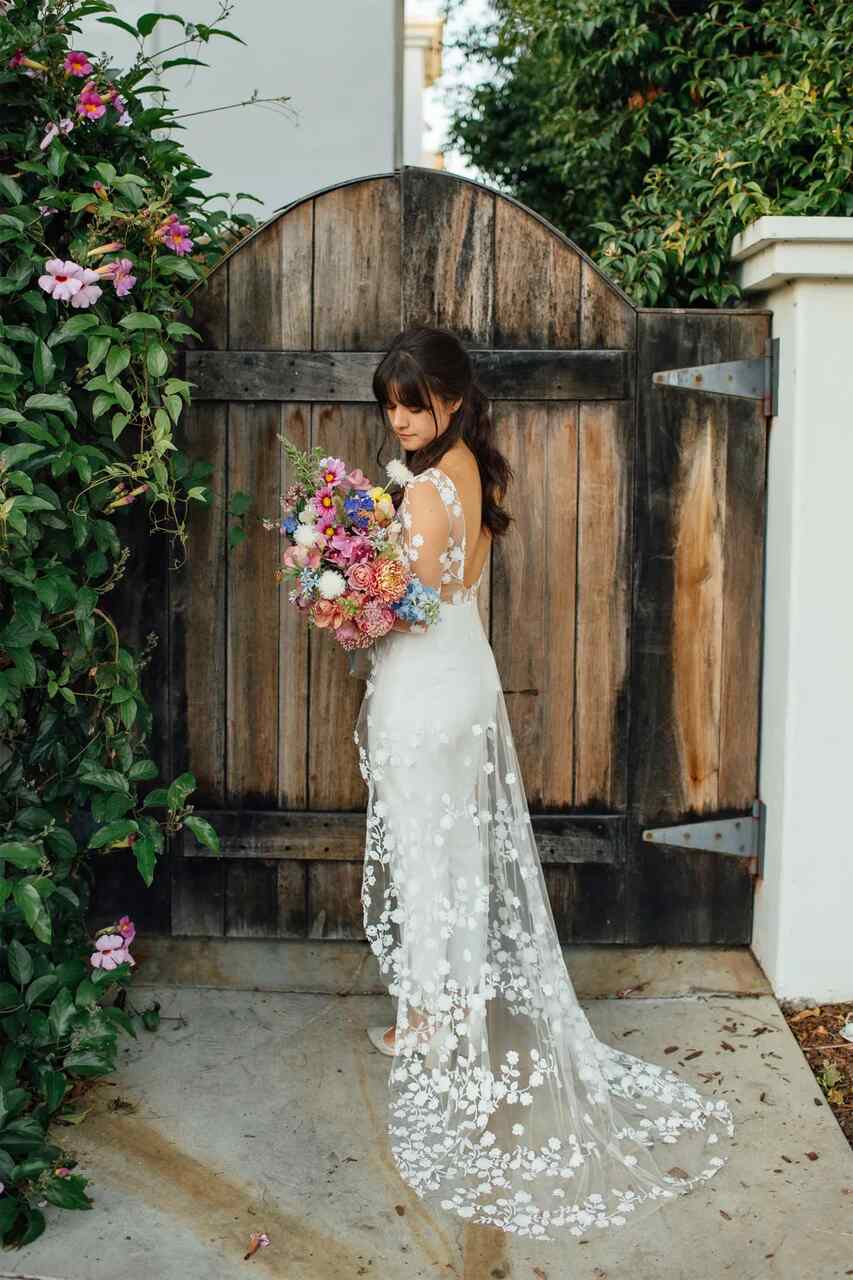 Uma noiva posando ao lado de um portão rústico, vestindo um vestido com detalhes florais e segurando um buquê colorido.