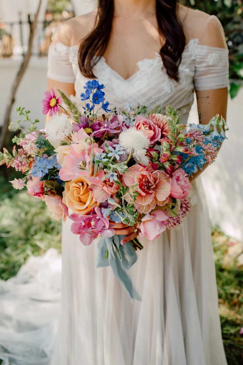 Uma noiva segurando um buquê multicolorido de flores em tons de rosa, azul e laranja