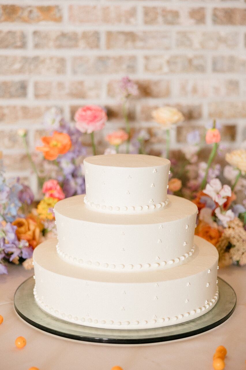 Bolo de casamento clássico de três andares, com cobertura branca e detalhes delicados de pequenas pérolas ao redor de cada camada. O bolo está em um suporte espelhado, com flores coloridas em tons pastel decorando o fundo