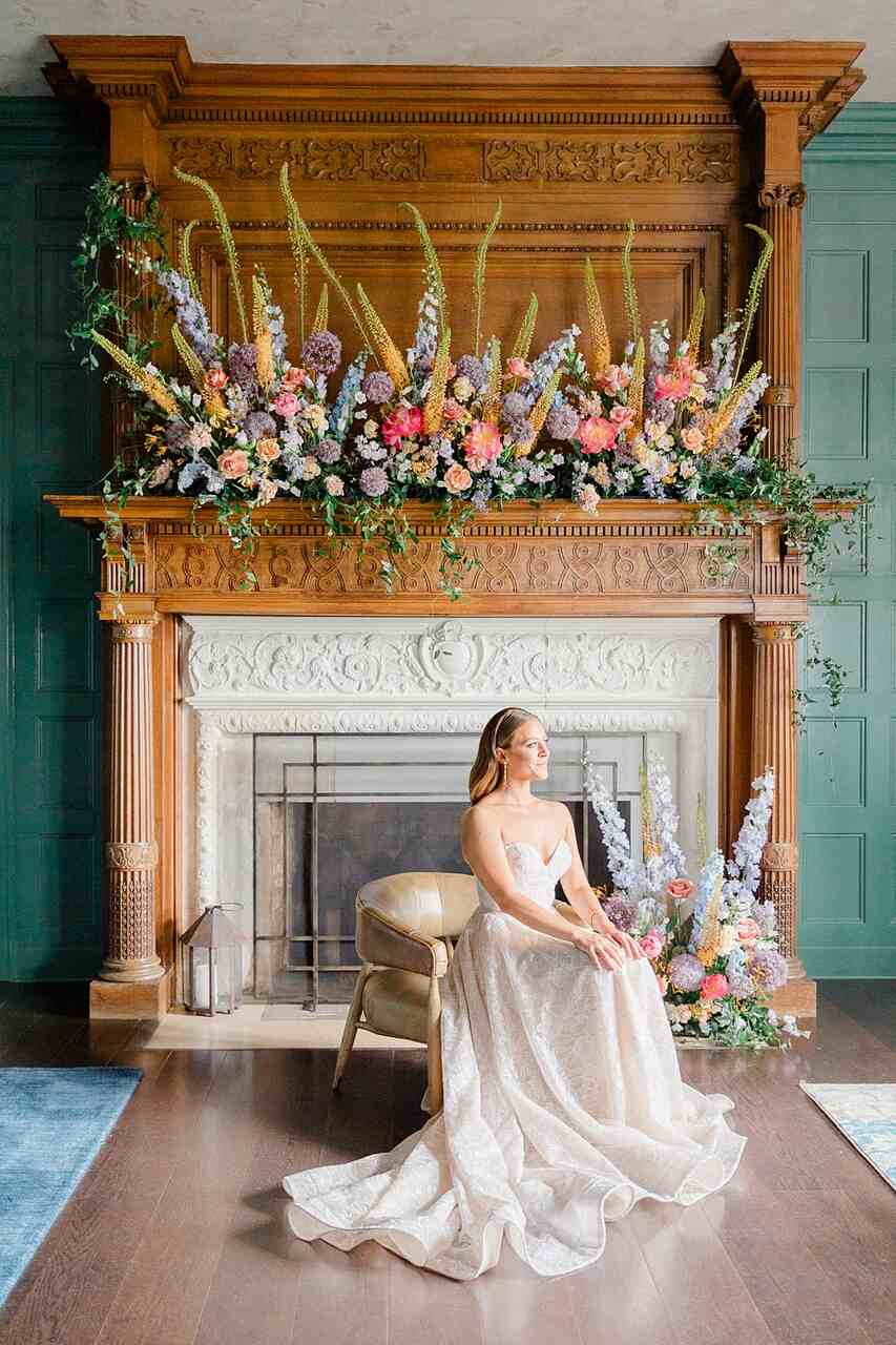 Noiva sentada em frente a uma lareira ornamentada com uma exuberante decoração floral em tons pastel