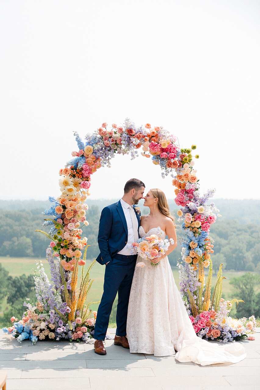 Casal de noivos emoldurado por um arco de flores em tons pastel, celebrando o amor em um cenário ao ar livre.