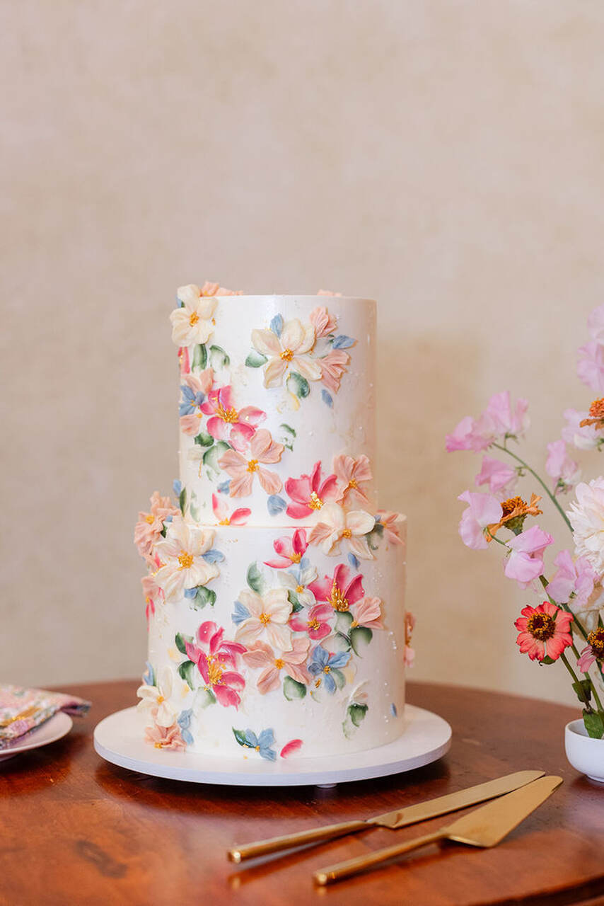 Bolo de casamento de dois andares com acabamento branco e decoração floral pintada à mão, incluindo flores coloridas em tons pastel, sobre uma mesa de madeira com espátulas douradas ao lado.