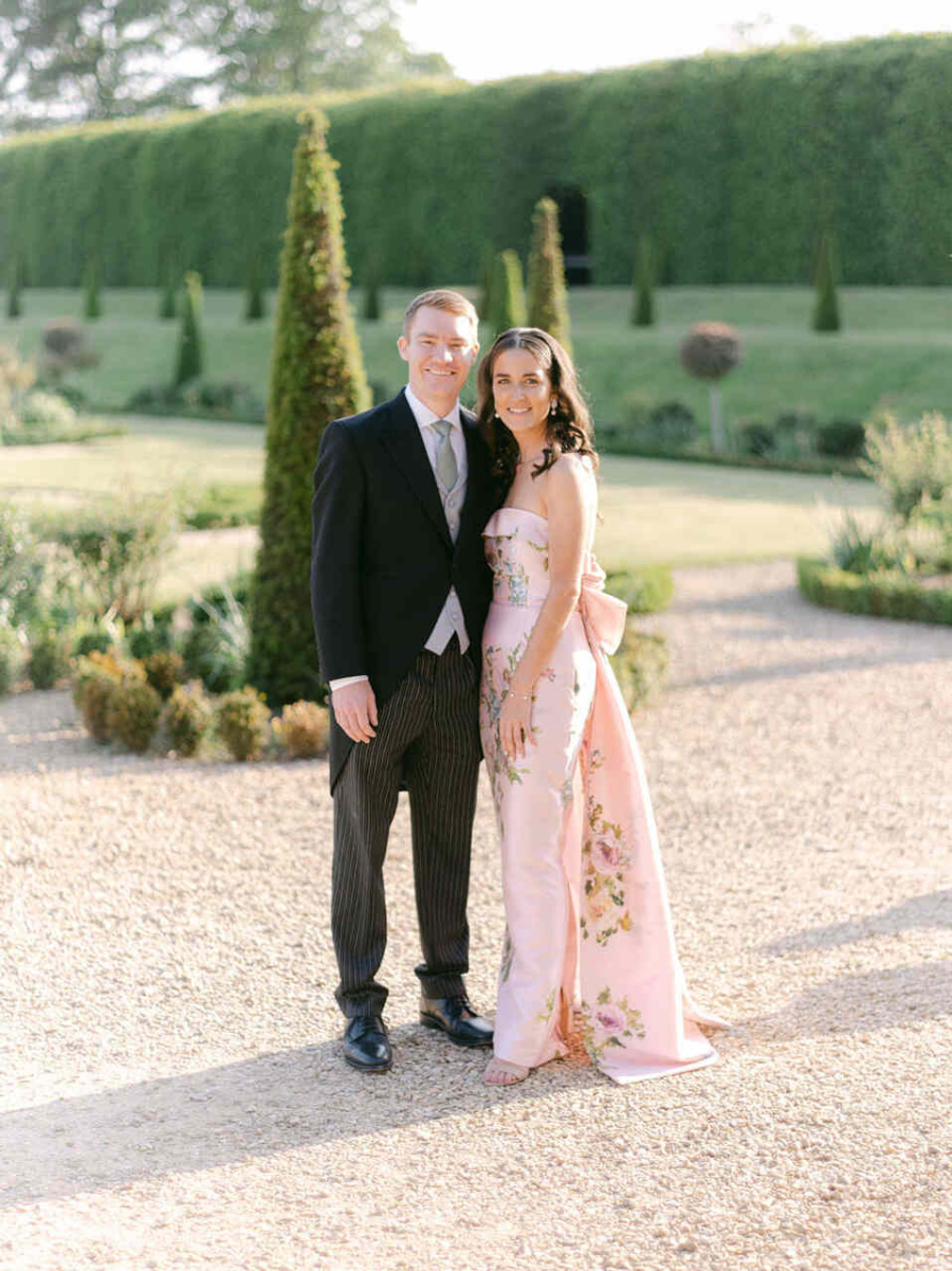 Casal em cenário de jardim; homem de fraque e mulher com vestido rosa claro floral com laço nas costas.