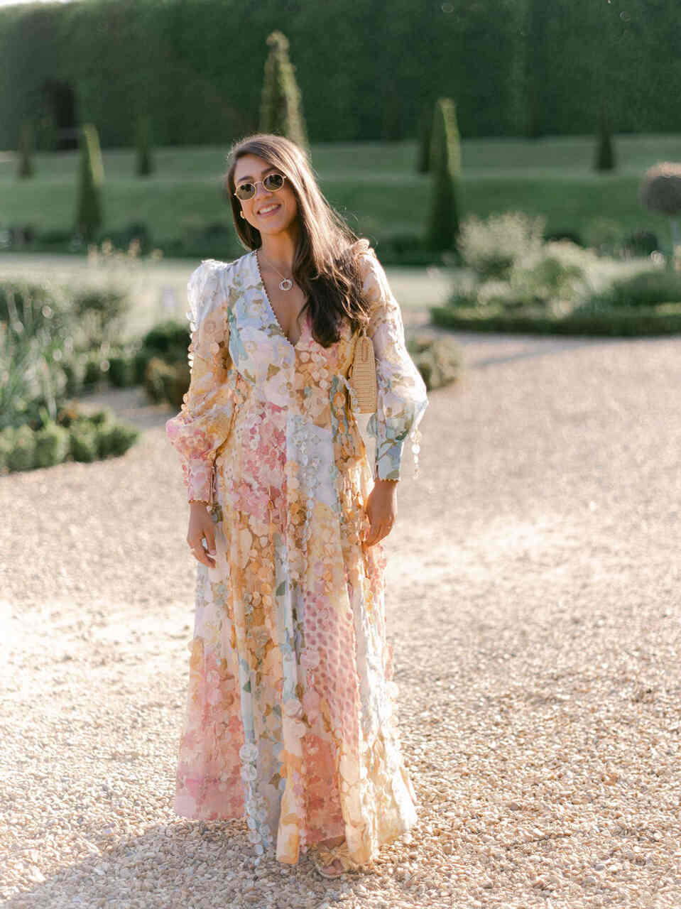 Mulher em jardim ao ar livre, usando vestido floral longo com mangas bufantes, óculos de sol e colar discreto.