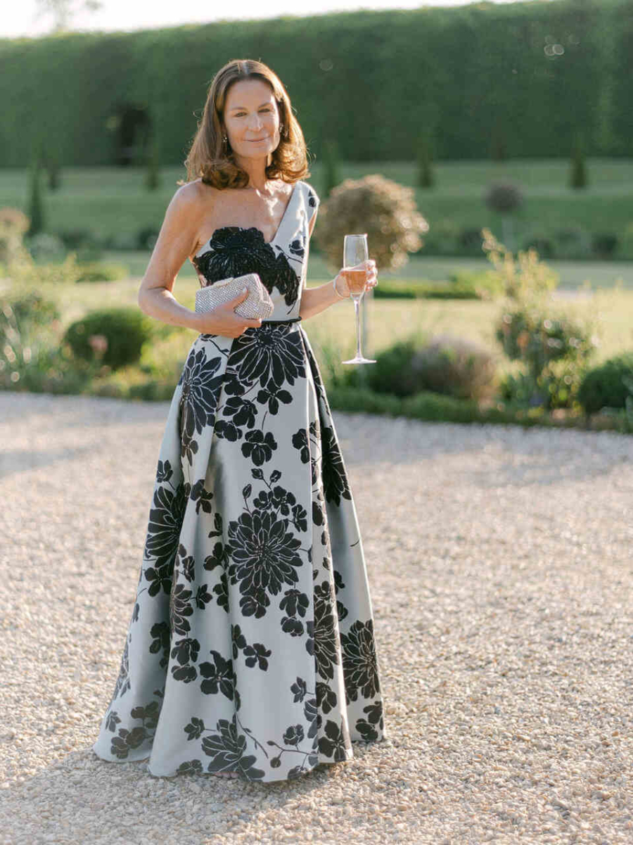 Mulher elegante em cenário de jardim, usando vestido longo branco com estampas florais pretas, segurando uma taça de champanhe e uma clutch prateada.