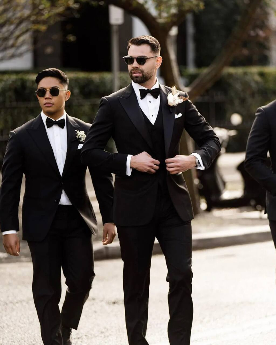 Três homens vestidos com smokings pretos caminhando em ambiente urbano