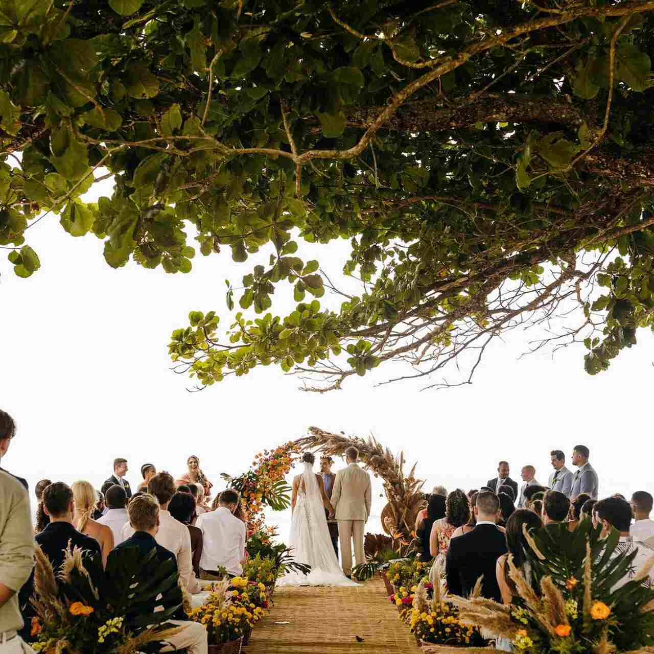 Casamento ao ar livre na praia, com uma cerimônia sob um arco floral, rodeada por convidados e árvores, criando um cenário natural e acolhedor.