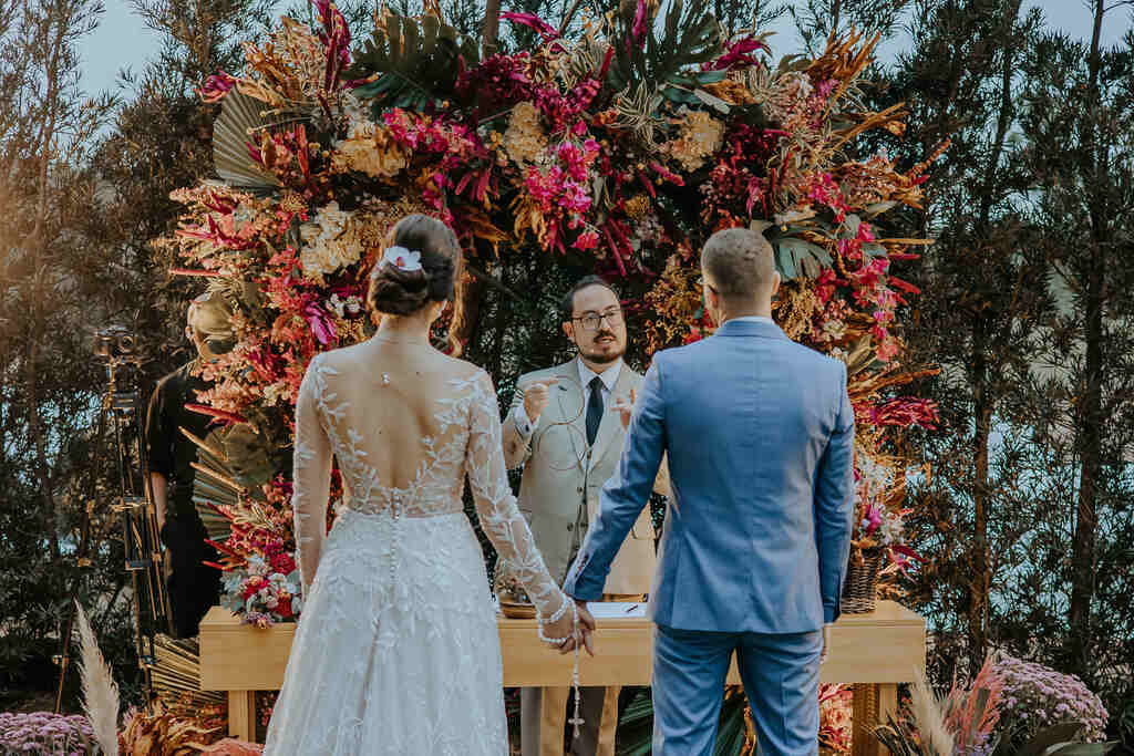 Casamento ao ar livre, com noivos de costas segurando as mãos, em frente a um celebrante. Ao fundo, uma exuberante decoração floral em tons de rosa e laranja.