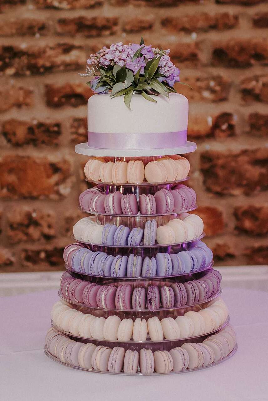 Torre de macarons em tons lilás e creme com um pequeno bolo branco no topo.