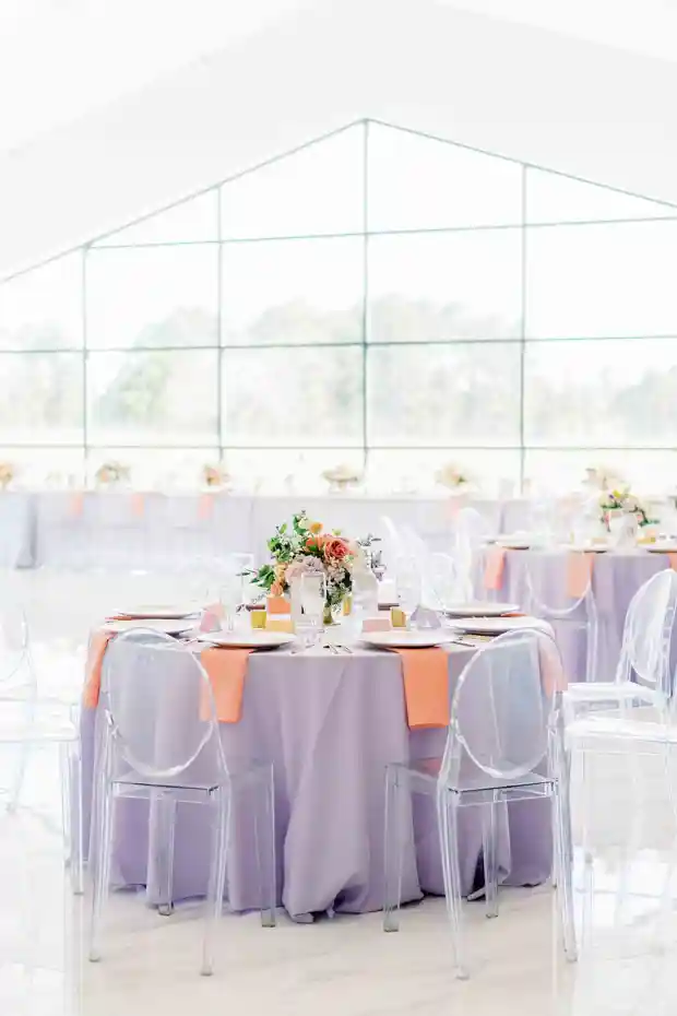 Mesa de casamento decorada com toalha lilás e cadeiras transparentes, com arranjo floral central