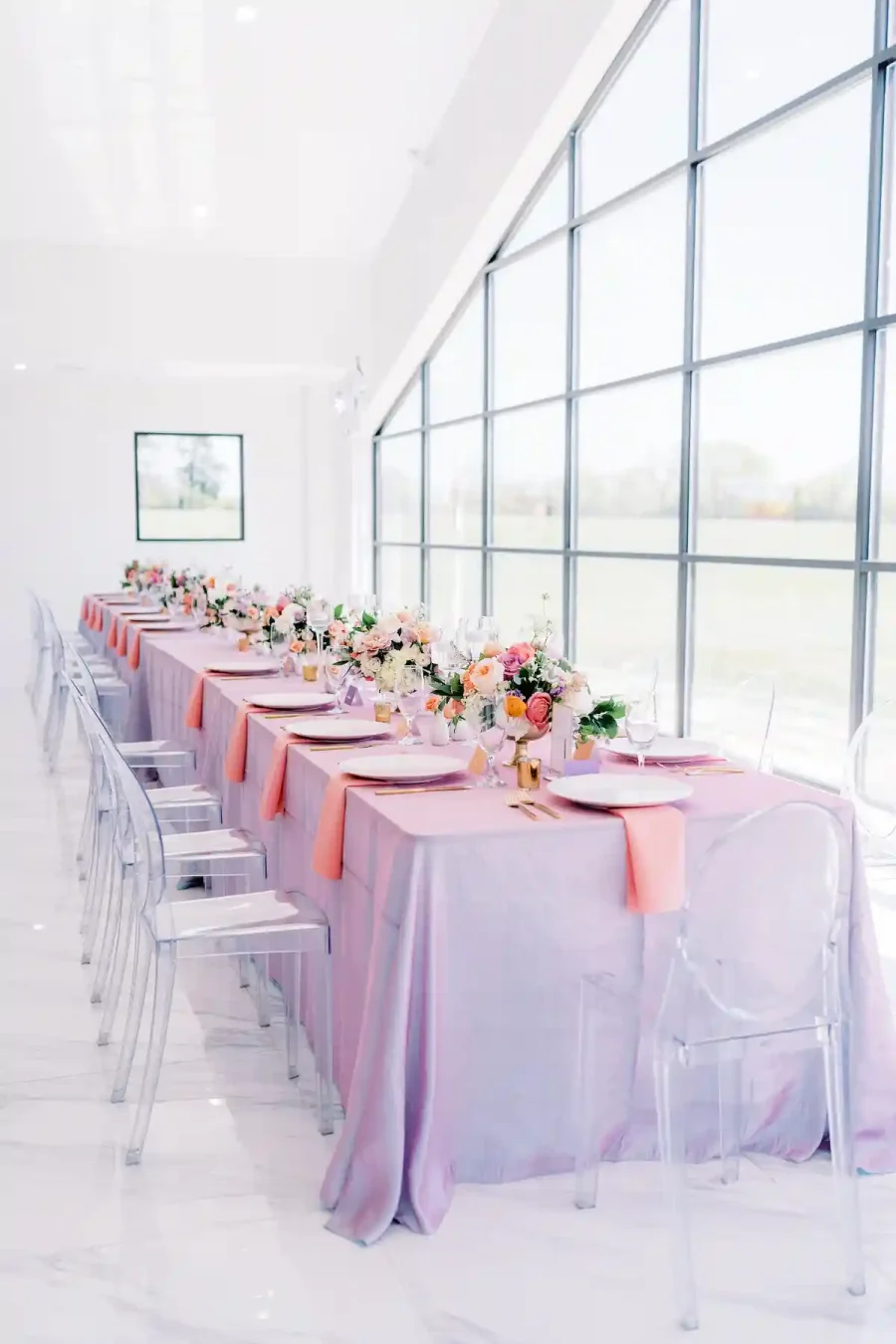 Mesa de casamento decorada com toalhas lilás, arranjos florais coloridos e cadeiras transparentes