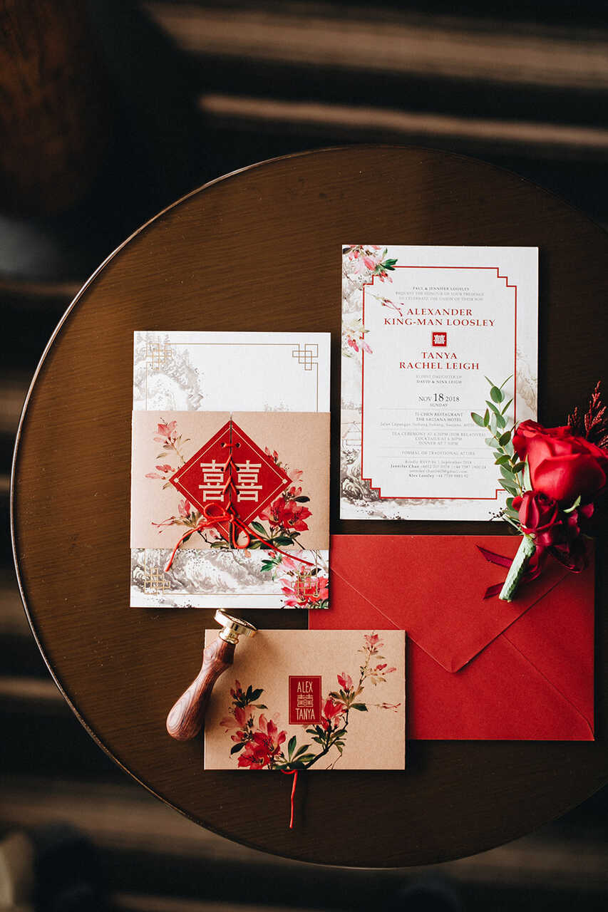 Imagem de convites de casamento com design inspirado em elementos tradicionais chineses, incluindo uma faixa vermelha com símbolos, flores e um carimbo. Os convites estão dispostos sobre uma mesa de madeira, com flores vermelhas e um envelope.