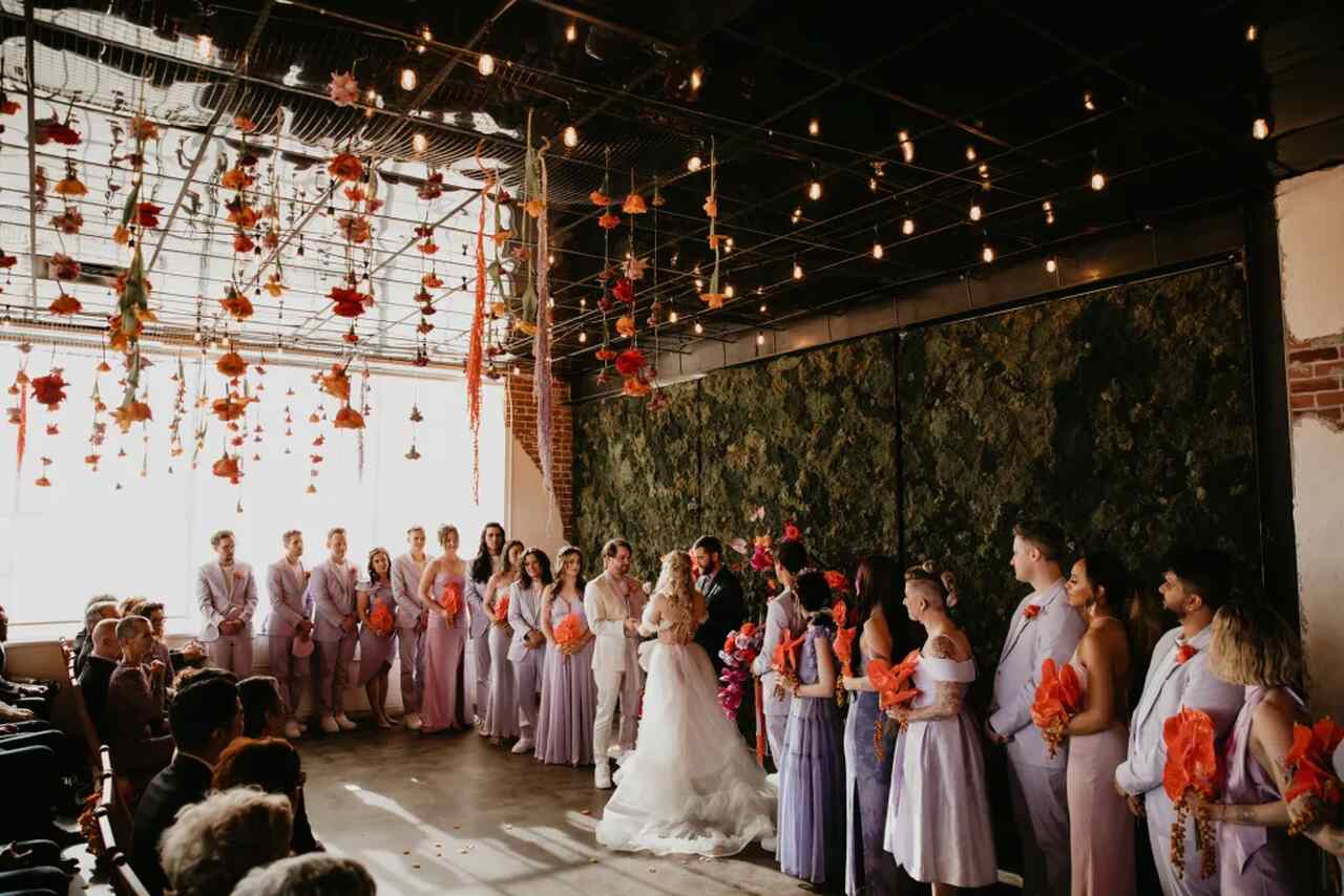 Noivos no altar, com casais de padrinhos ao lado, todos trajados com roupas na cor lilás.