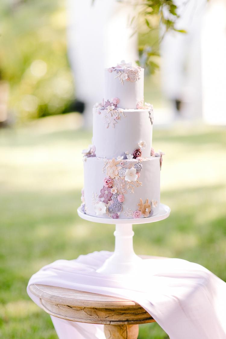 Bolo de casamento de três andares com acabamento em lilás, decorado com flores delicadas em tons de lavanda e rosa, posicionado em um ambiente ao ar livre.