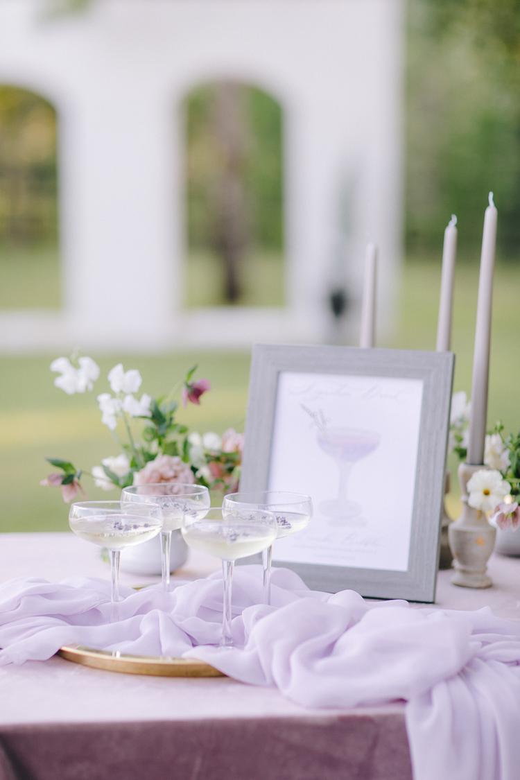 Configuração elegante de mesa com toalha lilás, quatro taças de drinks rodeadas por arranjos florais suaves e velas brancas.