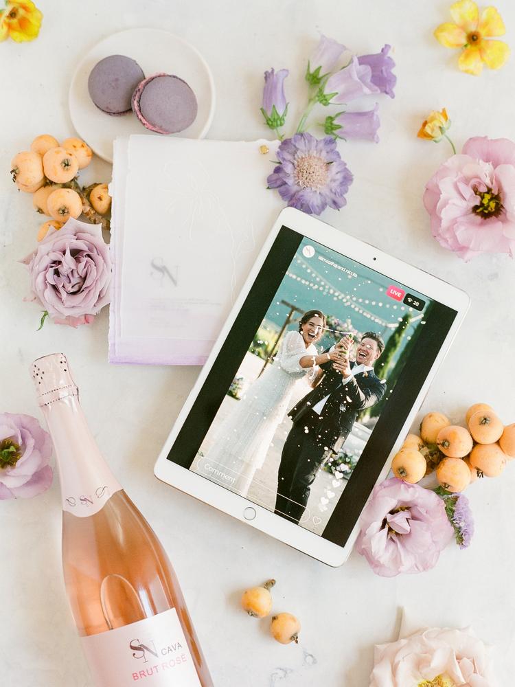 Mesa com uma garrafa de espumante rosé ao lado de flores lilás e rosa, uma tela com foto de casamento no tablet e macarons lilás