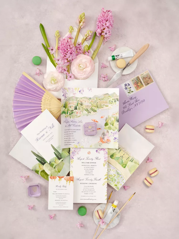 Conjunto de convites de casamento com design artístico e floral, com flores frescas e macarons, acompanhado de acessórios como selos e pincéis, em tons suaves de lilás e rosa.