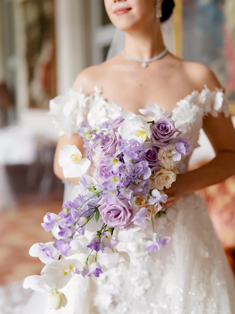 Buquê de noiva com flores lilases, brancas e orquídeas, complementando o vestido rendado.