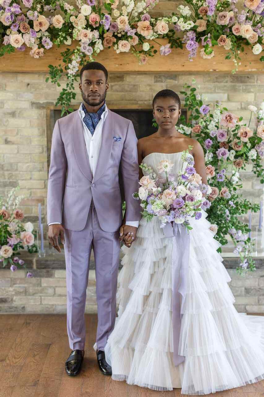 Casal posando em frente a uma lareira decorada com flores coloridas. O homem veste um terno lilás com lenço azul e a mulher usa um vestido branco com camadas e segura um buquê de flores em tons pastéis.