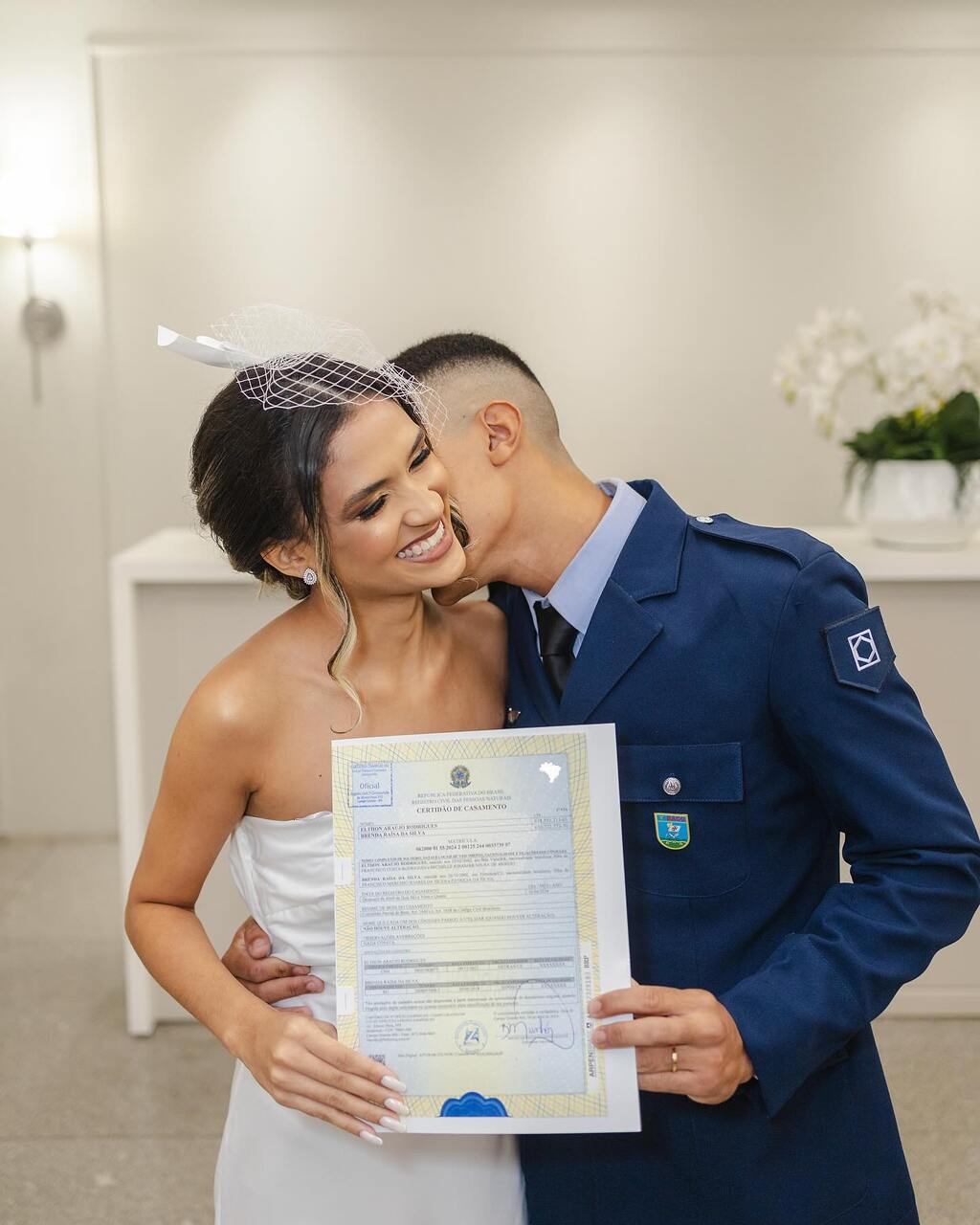 Noiva sorridente segurando a certidão de casamento enquanto o noivo, em uniforme militar, a abraça e demonstra carinho