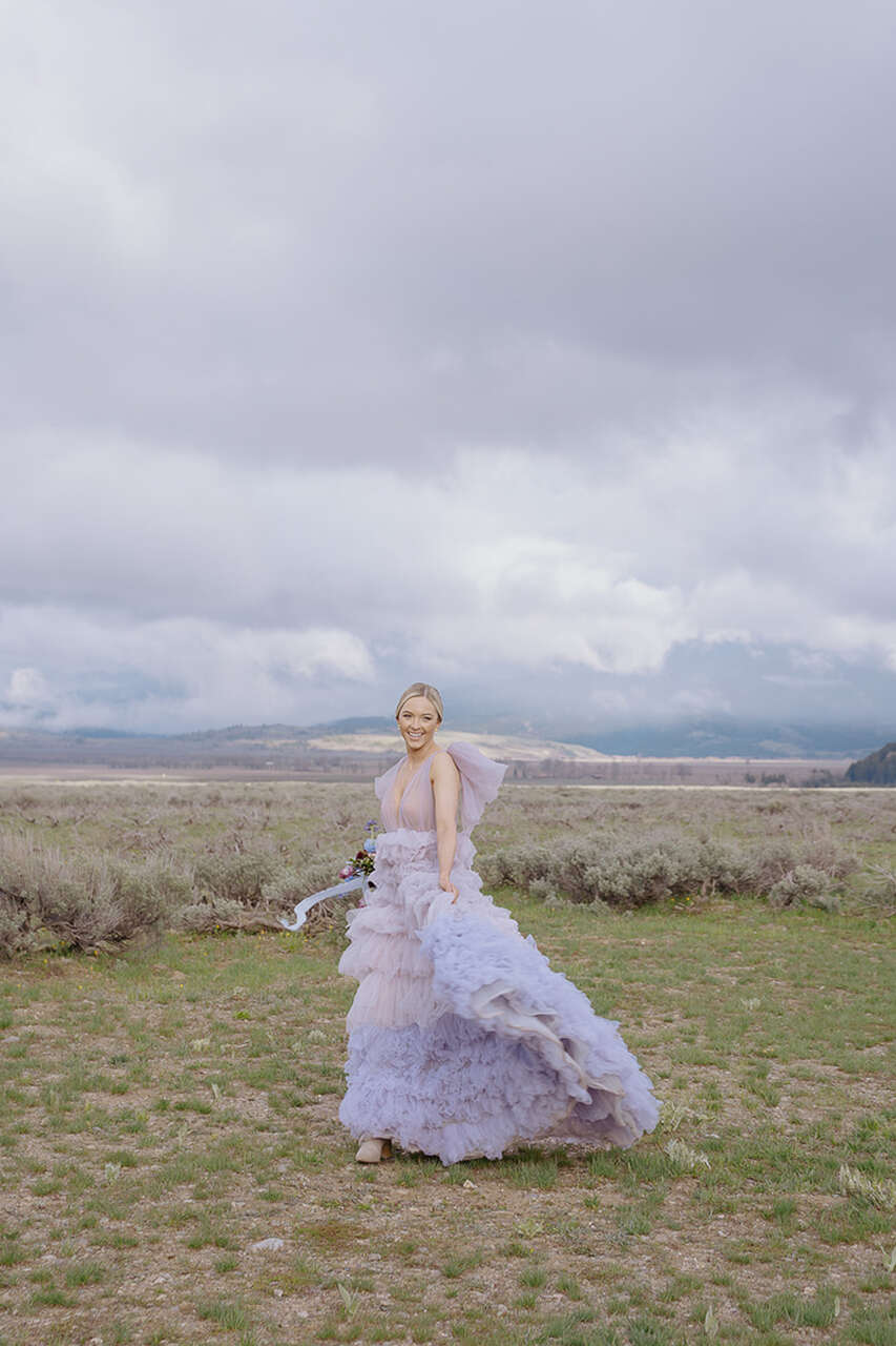 Noiva em campo aberto com vestido volumoso lilás, flutuando ao vento.