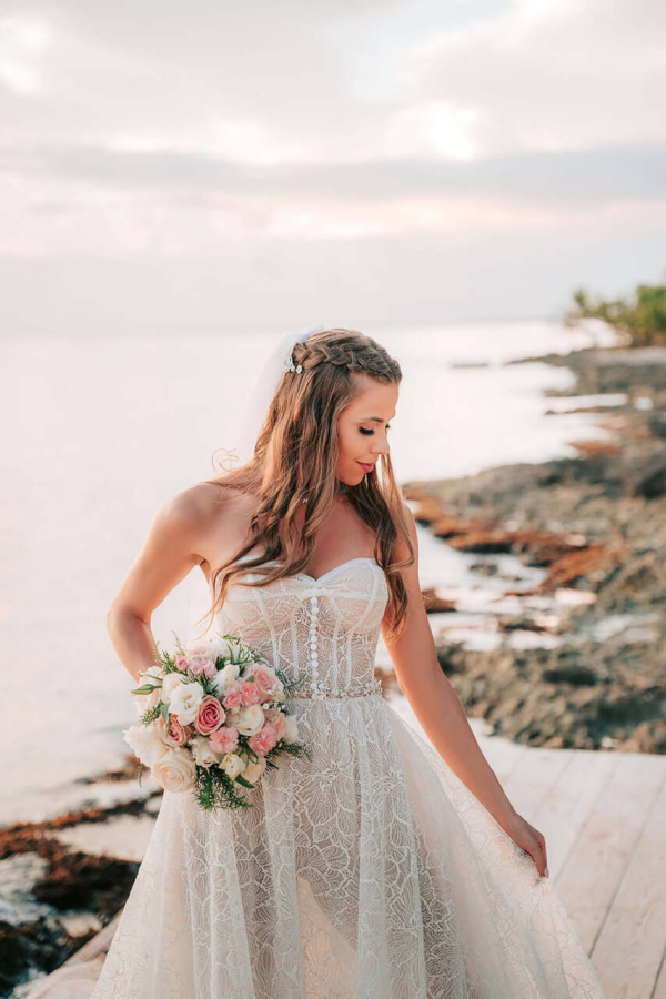capa post: Noiva usando um vestido de noiva delicado, com cabelo solto e uma trança, segurando um buquê de flores, enquanto caminha ao longo da praia com o mar ao fundo.