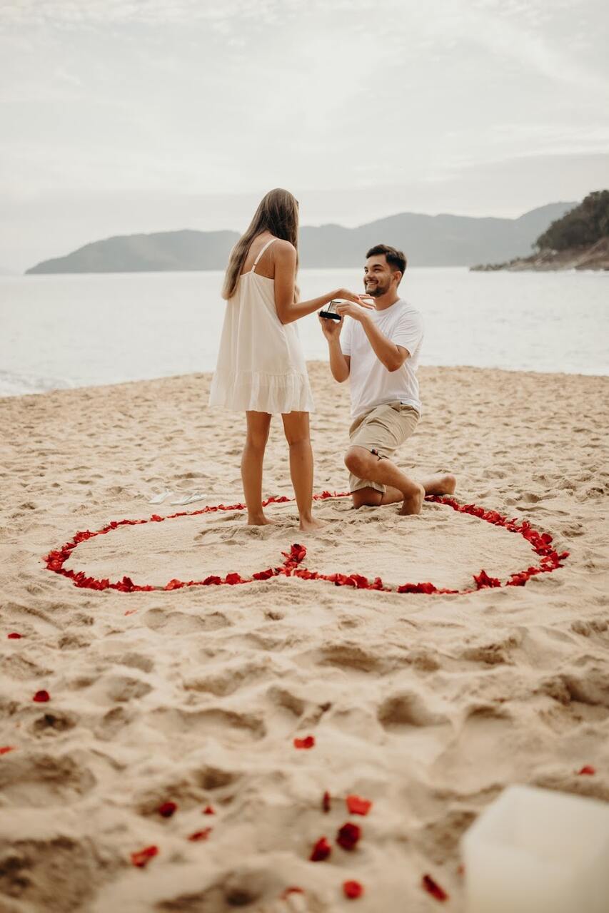 Homem fazendo o pedido de casamento na praia, de joelhos, enquanto a noiva aceita o anel, com pétalas de rosa formando um coração ao redor deles.