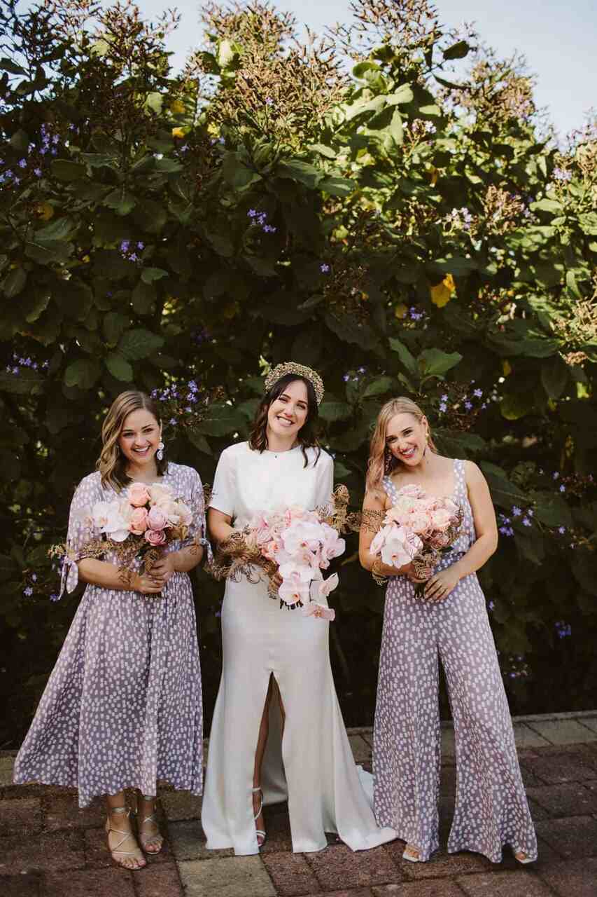 Noiva com amigas usando vestidos estampados lilases segurando buquês em um jardim exuberante.