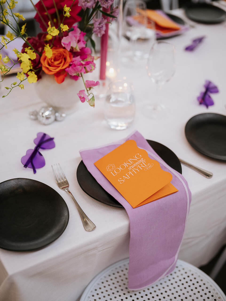 Mesa de jantar com toalha branca, talheres e pratos escuros, decorada com flores vibrantes e um convite laranja sobre um guardanapo lilás