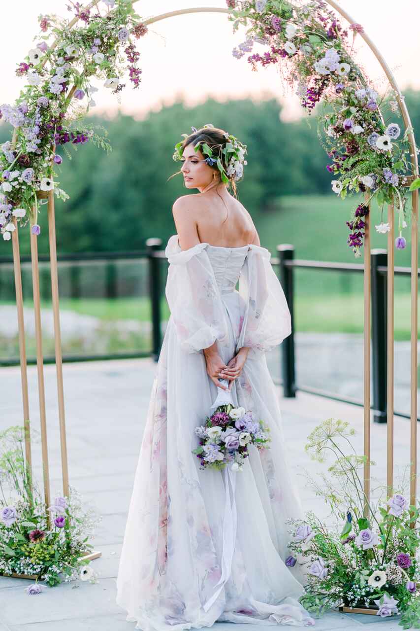 Noiva com vestido floral e buquê lilás, posicionada sob um arco decorado com flores.