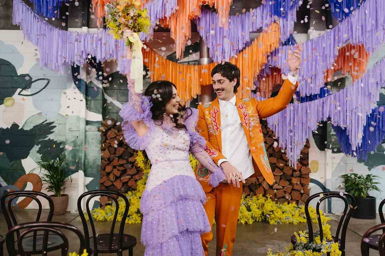 Casal celebrando em um ambiente vibrante e colorido, decorado com franjas roxas e laranja.