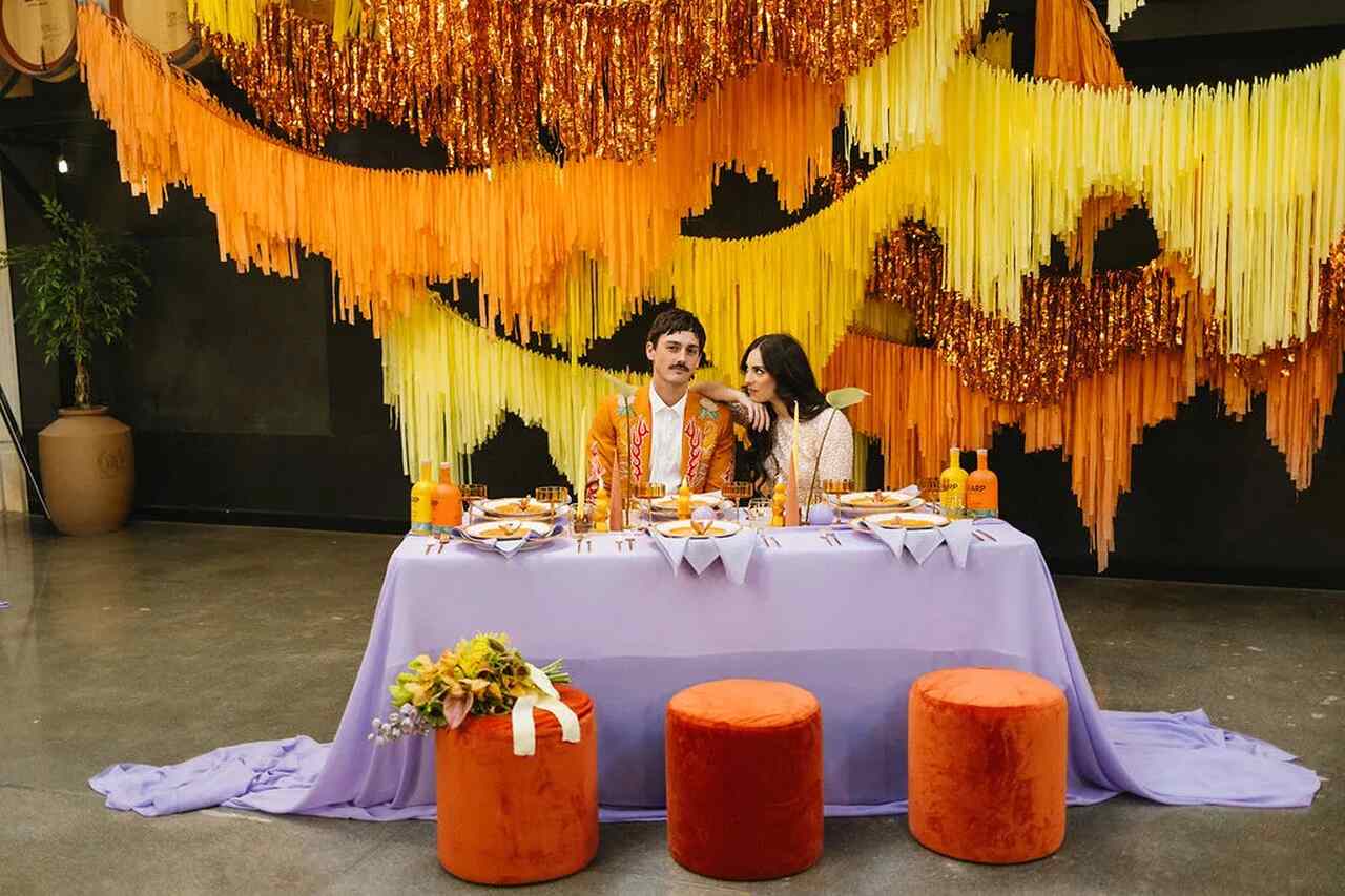 Mesa de casamento com tema boho, com toalha lilás, assentos laranja e decorações em amarelo e dourado