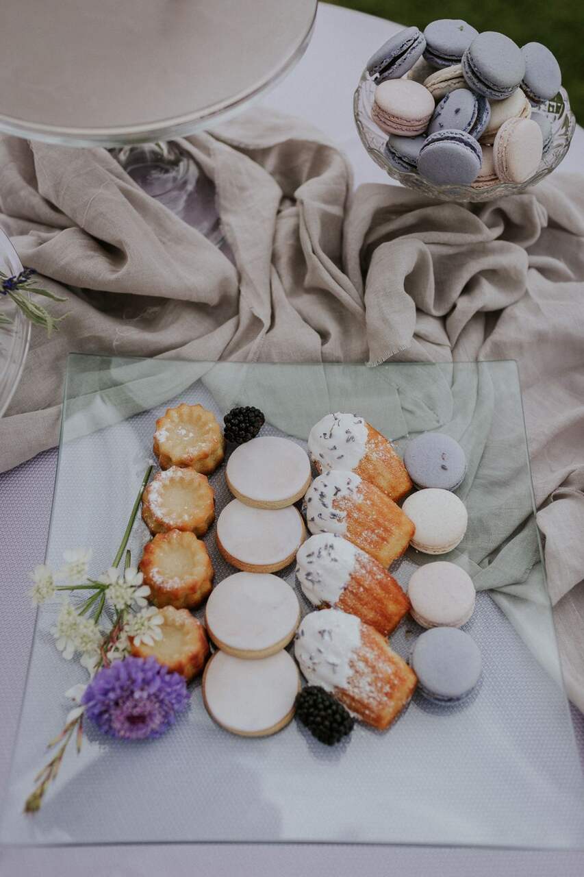 Bandeja de vidro com macarons, cookies e madeleines decoradas, cercada por flores frescas.