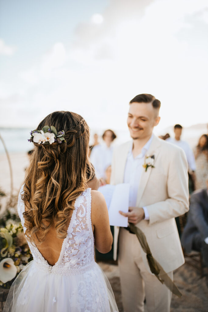 Noiva com vestido branco e flores no cabelo, durante a cerimônia de casamento na praia, enquanto troca votos com o noivo, que usa terno claro. Ao fundo, convidados observam o momento ao ar livre.