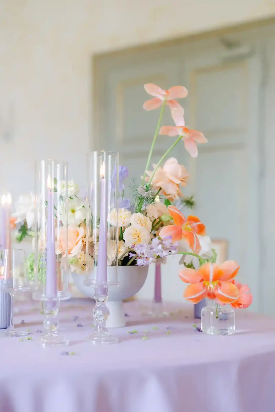 Centro de mesa de casamento com arranjo floral vibrante, combinando flores laranja, lilás e rosa, acompanhadas de velas altas em suportes elegantes