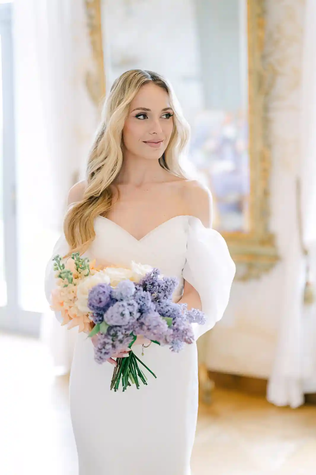 Noiva segurando um buquê de flores lilases e laranjas, com vestido branco de mangas bufantes.