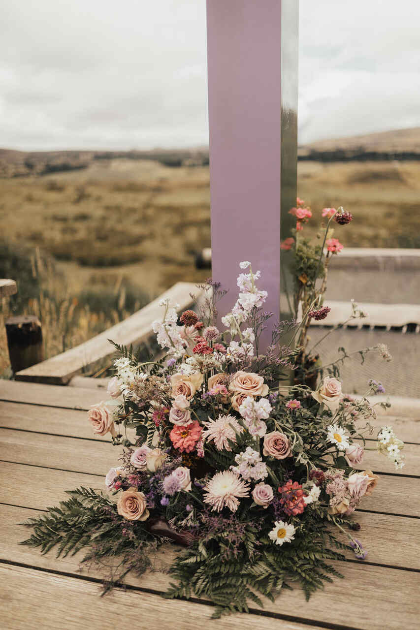 Arranjo floral com flores em tons de rosa, roxo e branco decorando o chão próximo a um pilar lilás, em um ambiente ao ar livre.