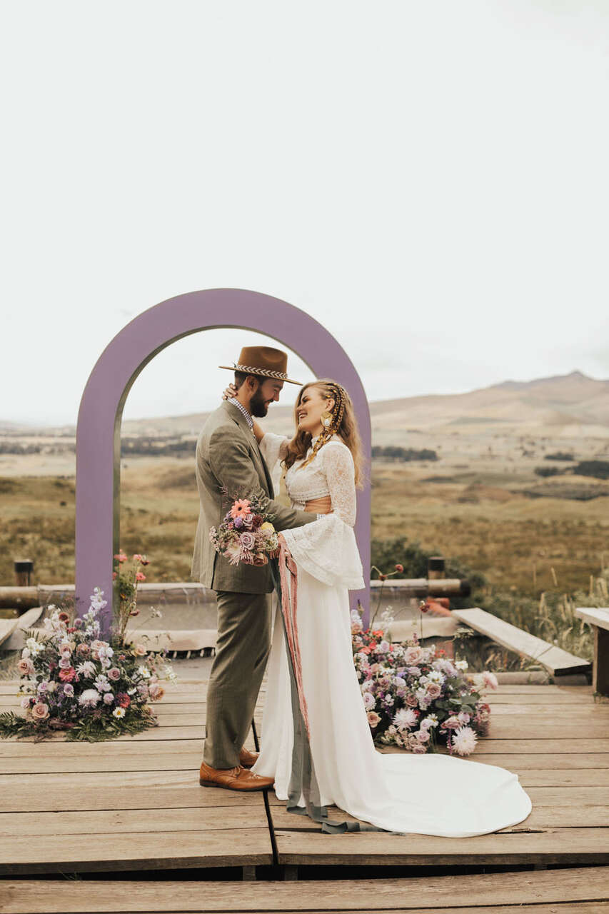 Casal ao ar livre em um cenário rústico. O homem veste terno cinza com chapéu e a mulher usa vestido branco com mangas rendadas, segurando um buquê. Ao fundo, um arco lilás decorado com flores.