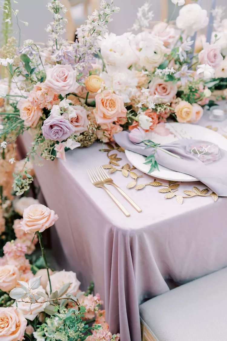 Mesa de jantar de casamento com toalha lilás, talheres dourados e arranjos florais delicados em tons de rosa, lilás e pêssego