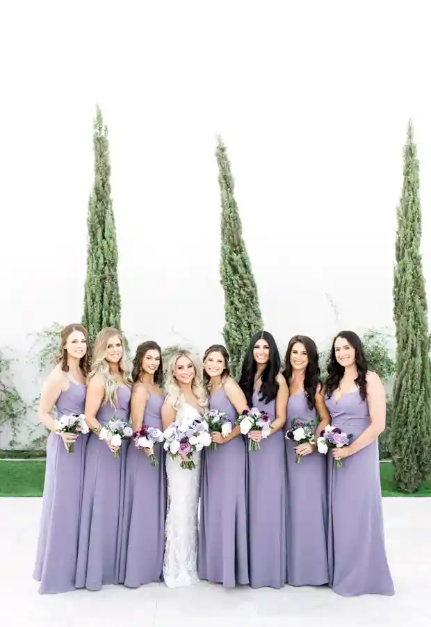 Noiva cercada por madrinhas em vestidos longos de lilás, segurando buquês de flores delicadas, com um fundo elegante.