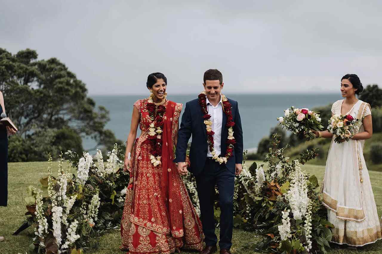 casamento ao ar livre com noivos caminhando de mãos dadas, sorrindo, enquanto decoradoras com flores em mãos acompanham. Ao fundo, o mar e uma paisagem verde.