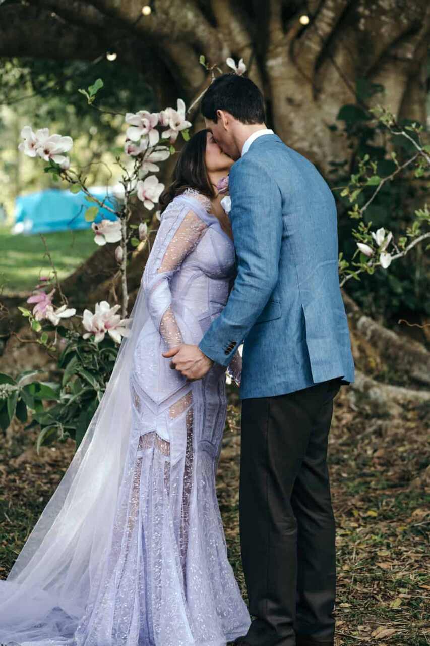 Casal trocando um beijo em um casamento ao ar livre, com um fundo de árvores e delicadas flores brancas e lilases decorando o cenário.