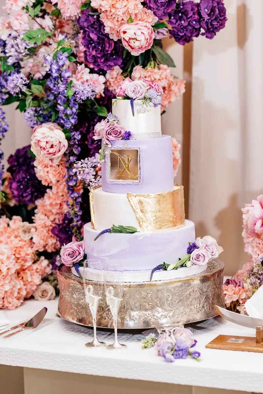 Bolo de casamento de três camadas decorado com flores lilases e dourado, rodeado por um fundo de flores em tons de roxo e rosa.