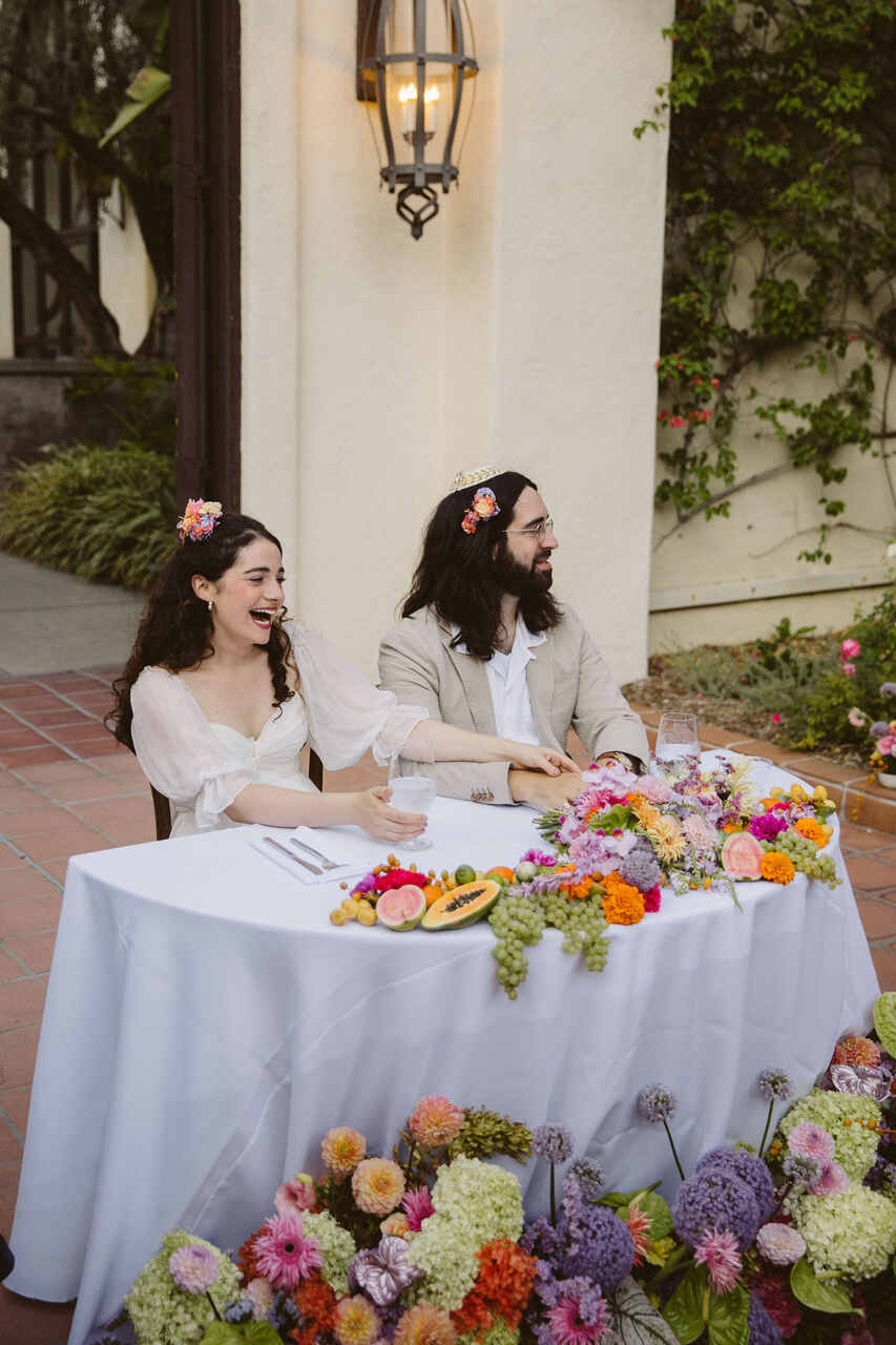 Casal sorrindo durante o jantar de casamento, com flores coloridas na mesa e tiaras florais em seus cabelos