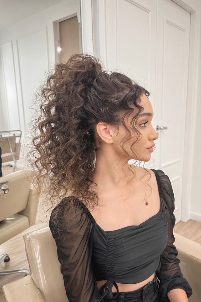 Mulher com cabelo cacheado, volumoso e preso parcialmente, com cachos soltos na parte inferior, com blusa preta 