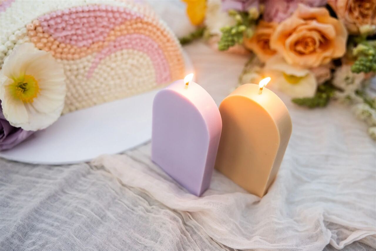 Velas decorativas em tons lilás e bege, acesas, com flores frescas ao fundo