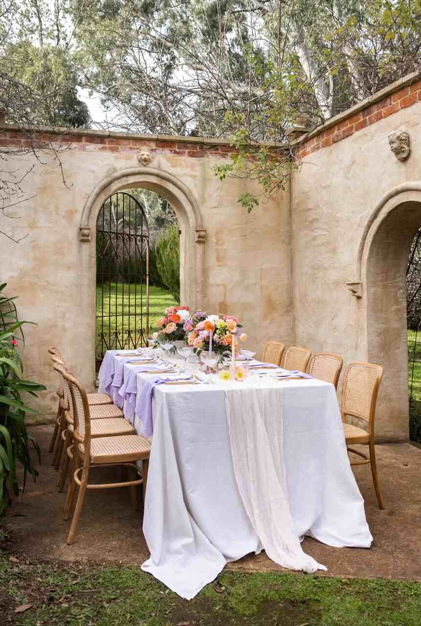 Mesa de casamento ao ar livre com toalha lilás e arranjo floral vibrante em tons de rosa e laranja, com uma vista de jardim