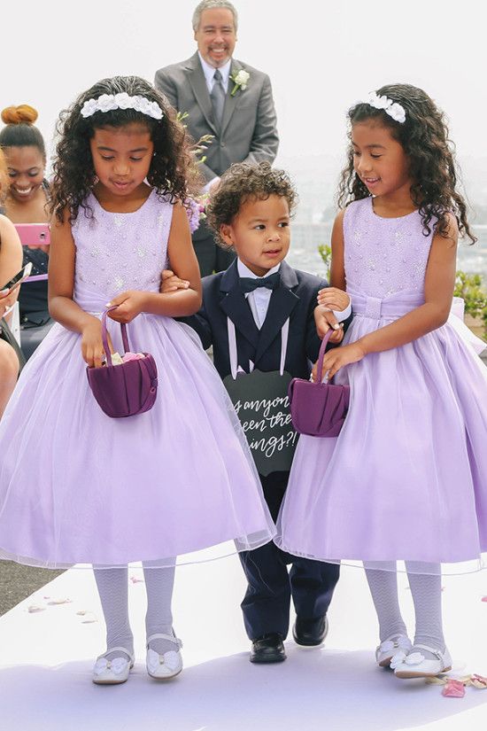 Duas daminhas com vestidos lilases segurando cestas e um pajem elegante de smoking com uma plaquinha divertida.