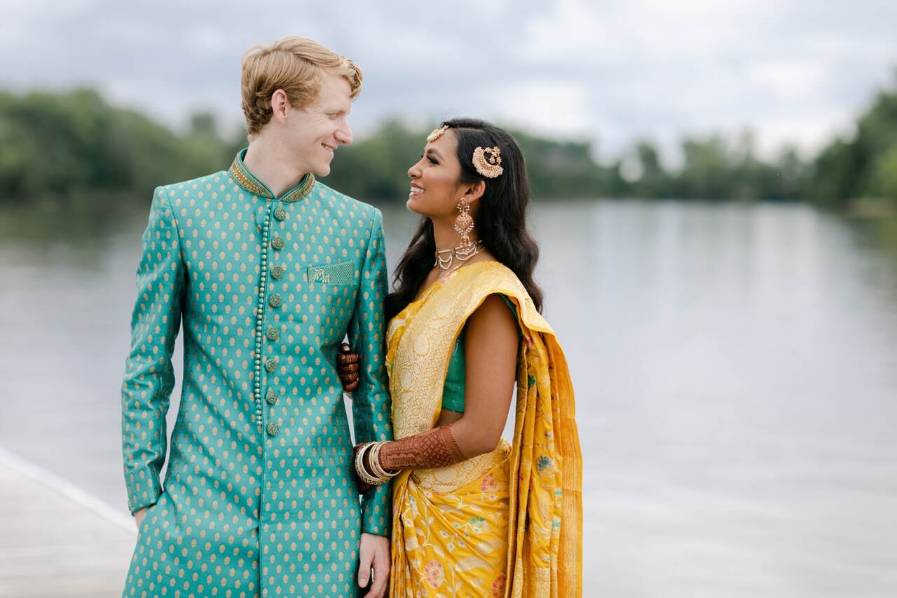 Noivos sorrindo um para o outro, vestidos com roupas tradicionais em tons de verde e dourado. A noiva usa um sari amarelo com detalhes dourados, enquanto o noivo veste um traje de festa indiano em verde com estampas douradas, em frente a um cenário ao ar livre com água ao fundo.