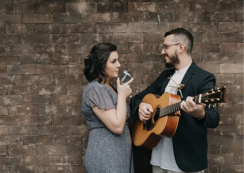 dupla de músicos, Mari e Gu, conhecidos por performances em cerimônias de casamento, posando em frente a uma parede de tijolos.